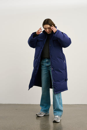 A model wearing a long navy blue puffer coat from MEOTINE, paired with a dark sweater and light blue jeans, striking a thoughtful pose.
