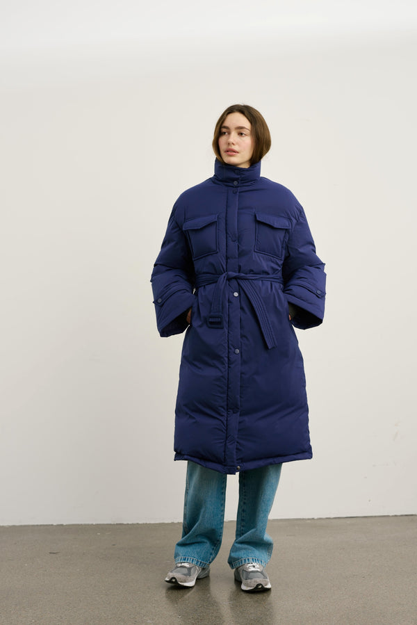 A model wears a long navy puffer coat with a belted waist and large pockets, paired with light blue jeans and sneakers. MEOTINE.