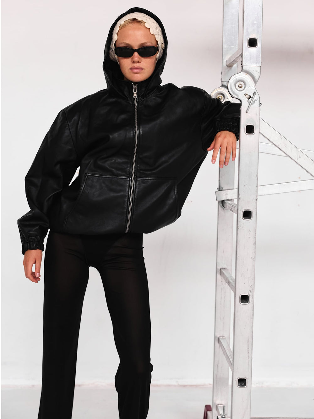 Stylish black leather jacket with a hood, featuring a zip closure and relaxed fit, paired with sleek sunglasses. MEOTINE.