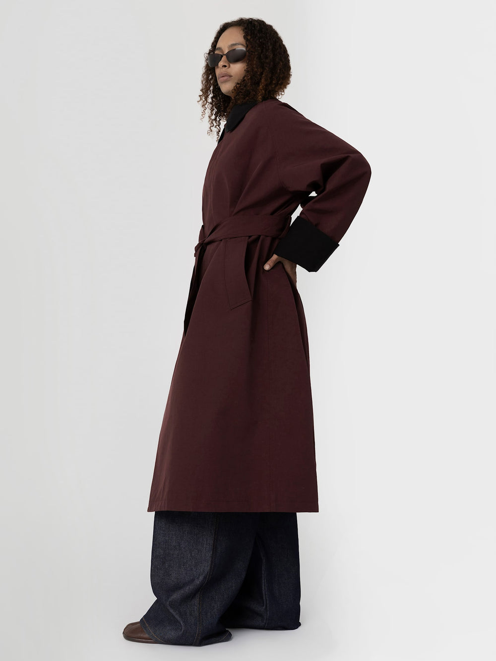 A model showcases a stylish burgundy coat with a tie waist and contrasting black cuffs, paired with wide-leg denim pants. MEOTINE.