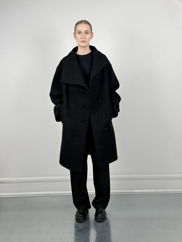 A model showcases a stylish black oversized coat by MEOTINE, paired with a sleek black outfit against a minimalist backdrop.